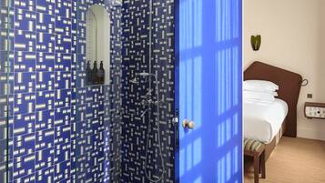 Un baño de hotel con un diseño de azulejos azules estampados, cerramiento de ducha de vidrio y una cama visible en la habitación contigua.