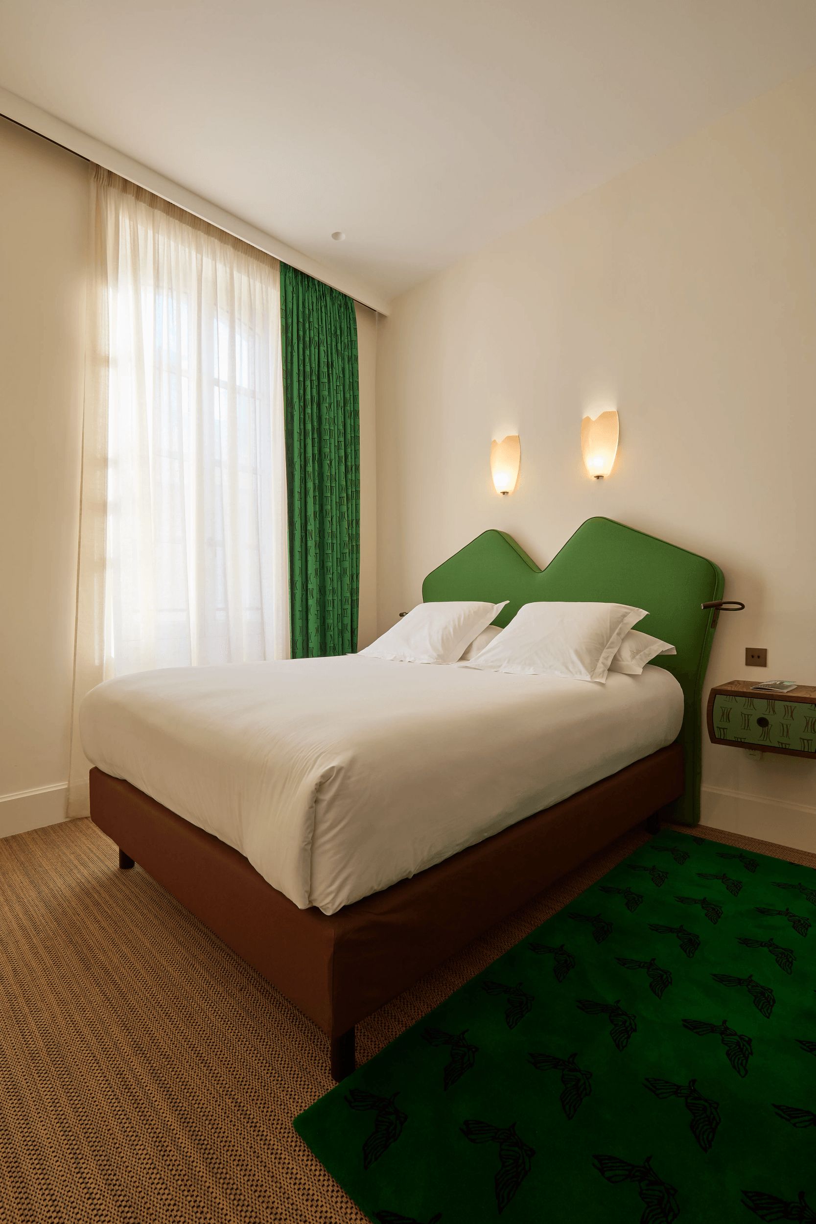 Une chambre d’hôtel avec un lit, une tête de lit verte, deux lampes fixées au mur, une fenêtre avec des rideaux légers et un tapis à motifs verts.