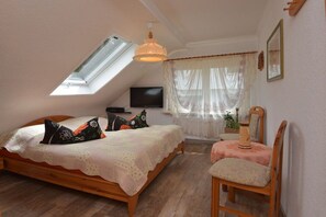 Ein Schlafzimmer mit schrägem Dach, einem Bett mit Häkeldecke, einem Sessel, einem Fernseher und einem Fenster mit durchsichtigen Vorhängen.