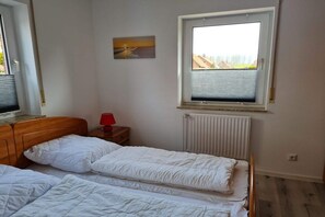 Quarto com duas camas, janela com persianas, radiador e uma pequena mesa com abajur vermelho.