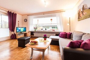 Ein Wohnzimmer mit Fernseher, einer Couch, einem Couchtisch mit Obst und einem Fenster mit Vorhängen.