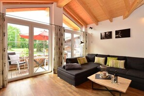 A modern living room with a black sofa, wooden flooring, and a glass door leading to an outdoor area with a red umbrella. - Holiday apartment with 1 bedroom (Fischen)