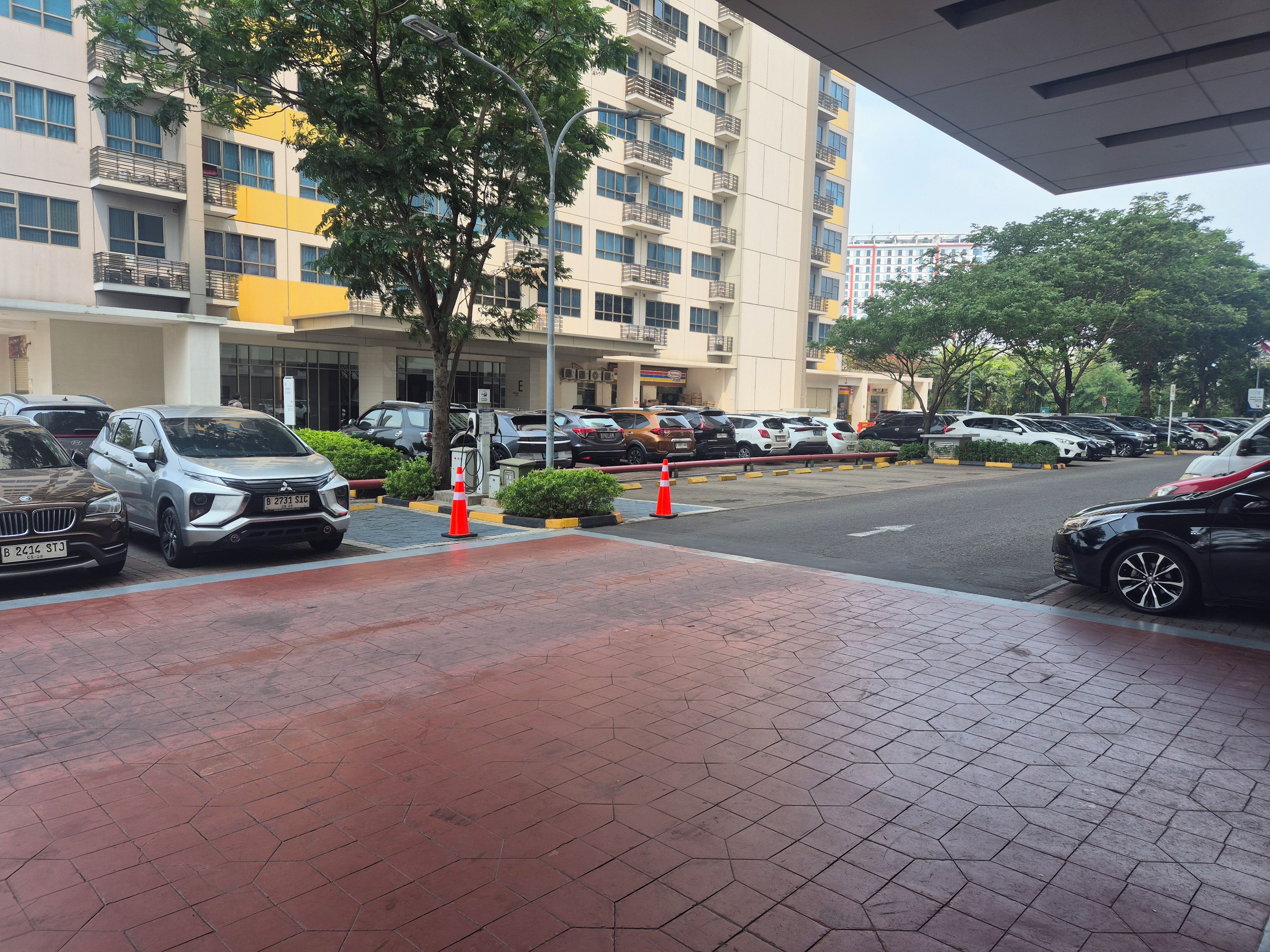 A parking area with multiple cars, a building with balconies, and a tree in the foreground.