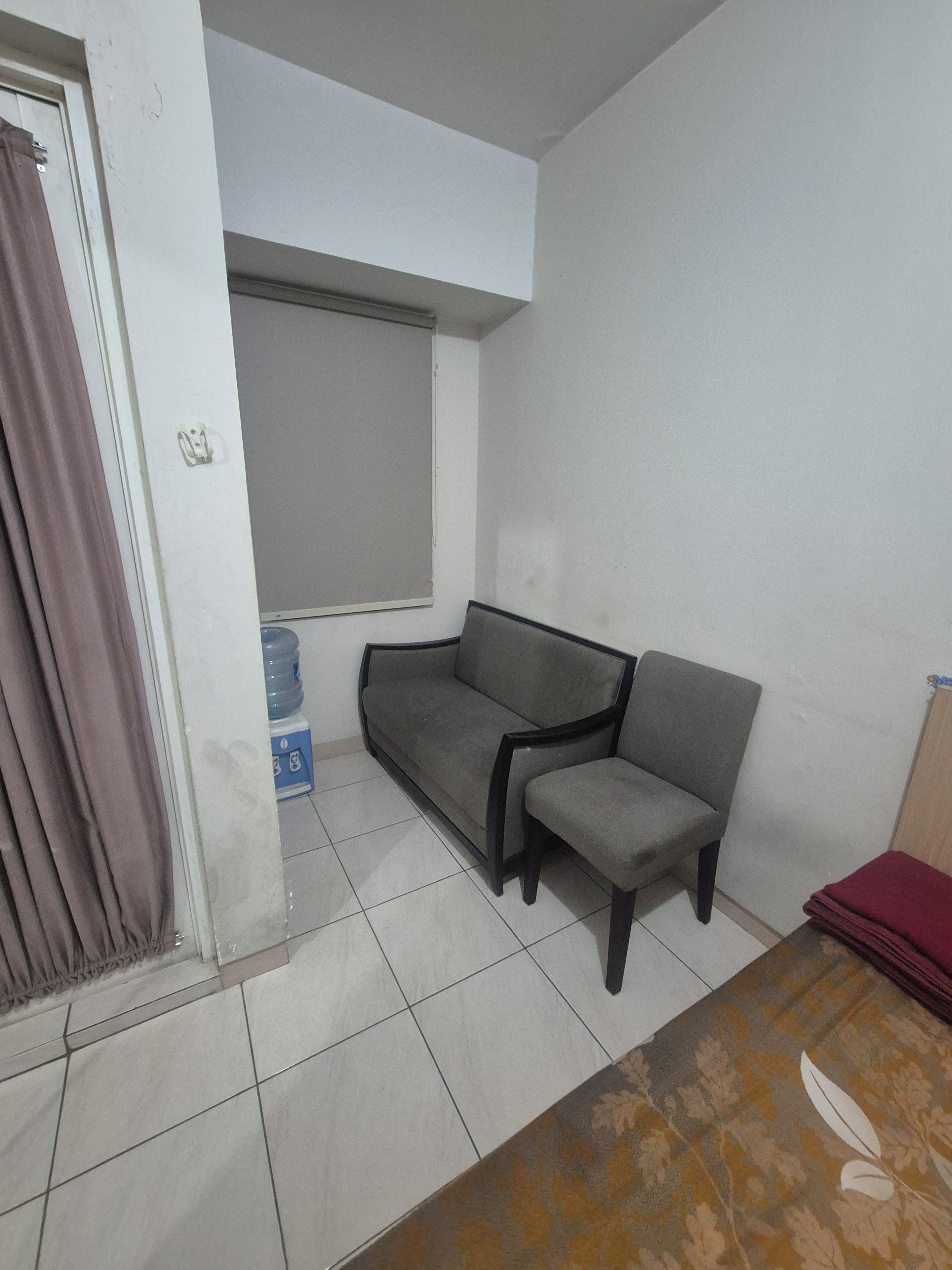 A room with a grey sofa, a small chair, a water dispenser, and a window with a blind.
