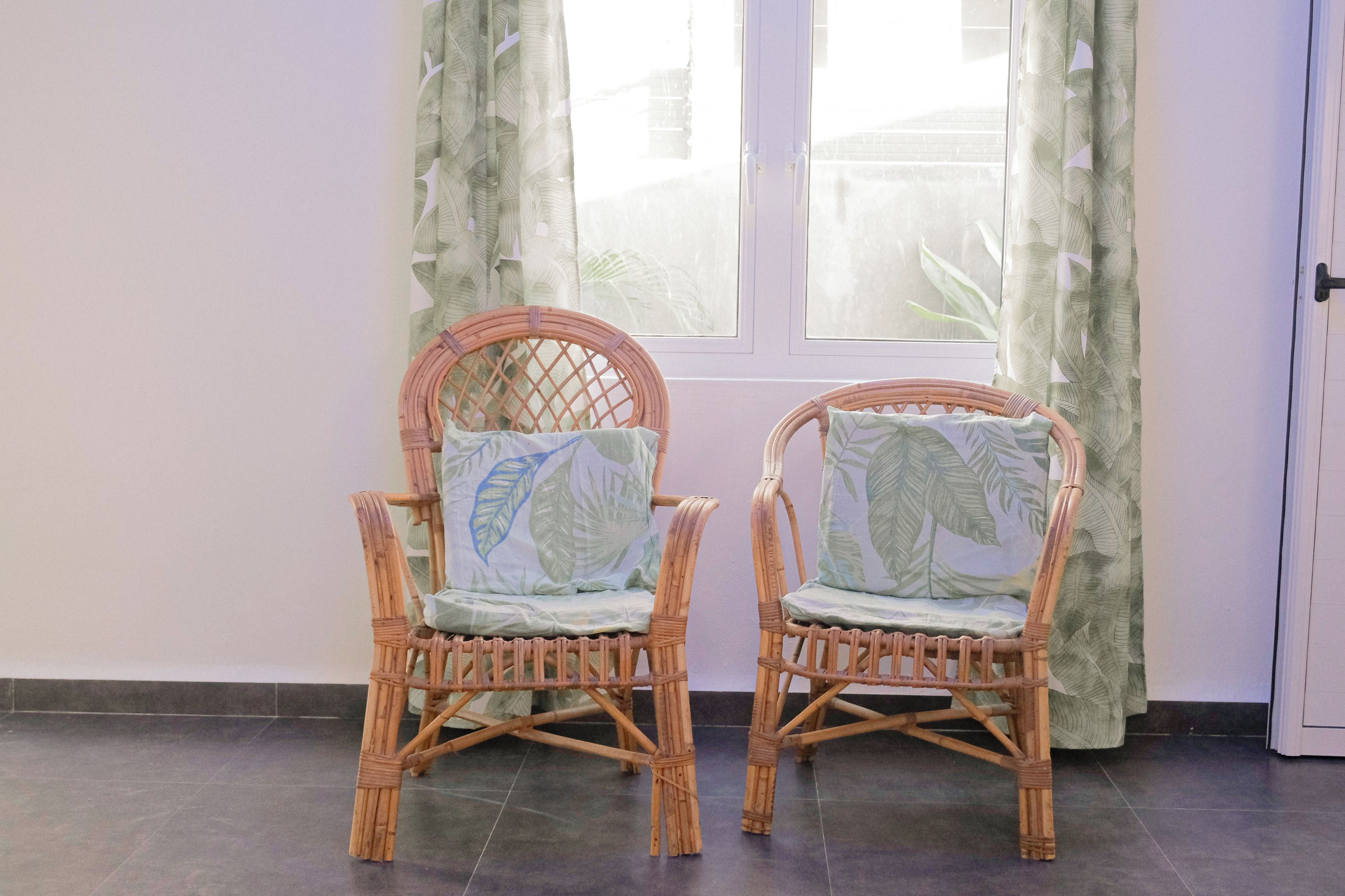 Two wicker chairs with leaf-patterned cushions in front of a window with curtains.