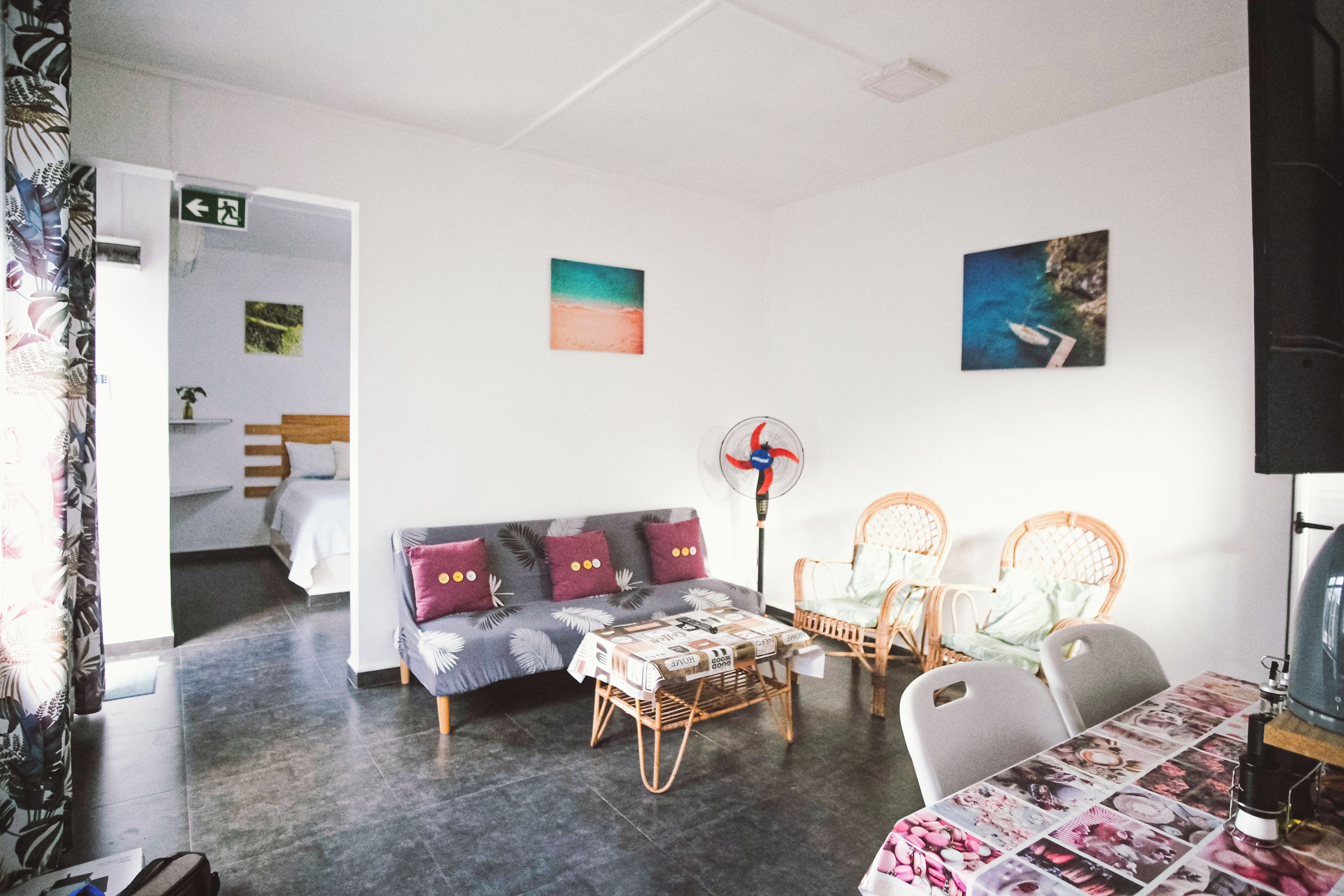 A room with a sofa, two chairs, a table, and a fan. There is a bed in the background.