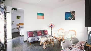 A room with a sofa, two chairs, a table, and a fan. There is a bed in the background.