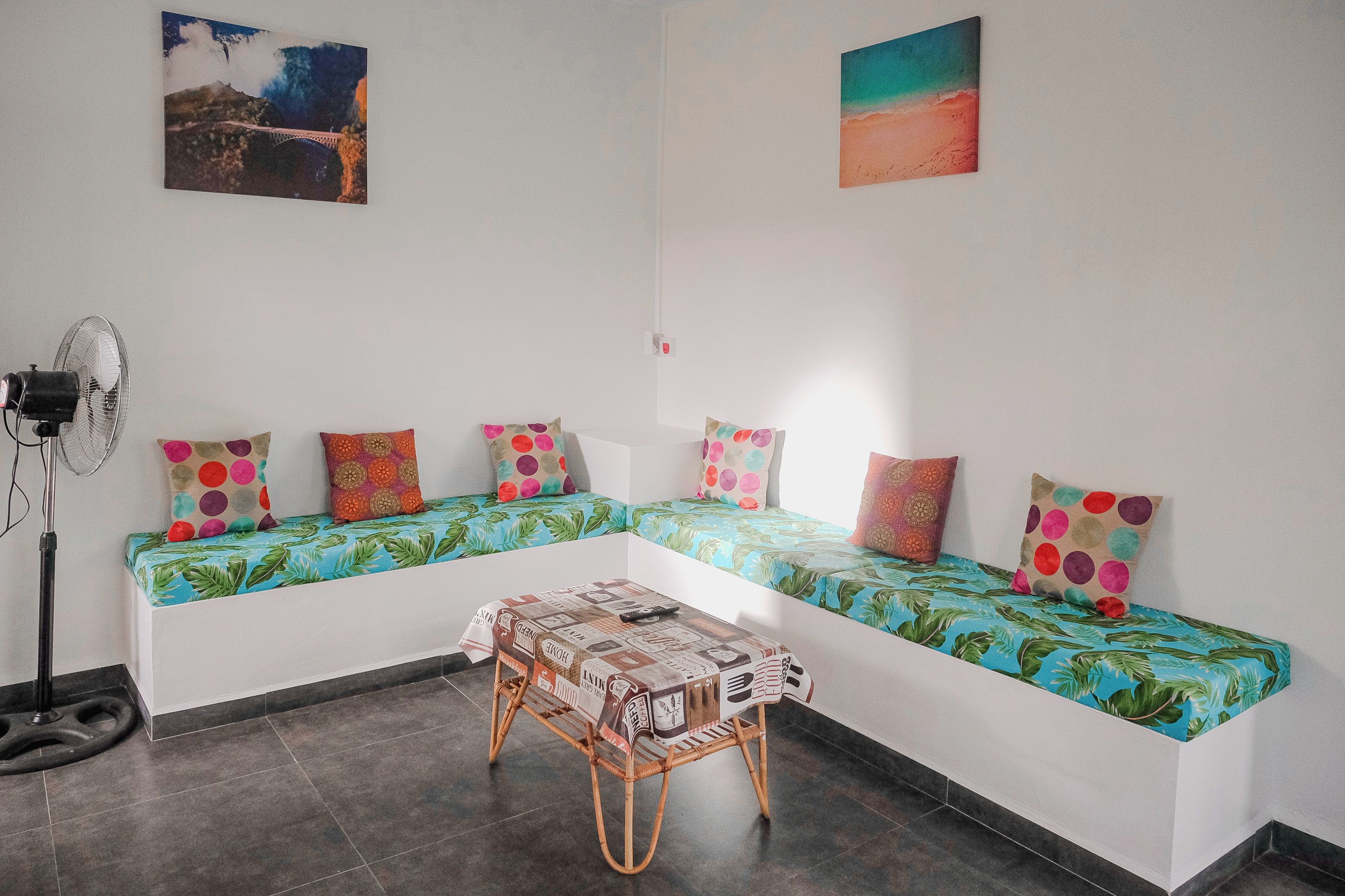 A room with a long white sofa with colorful cushions, a small table with a newspaper, and a fan.