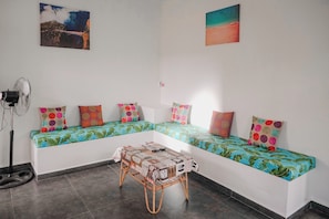 A room with a long white sofa with colorful cushions, a small table with a newspaper, and a fan.