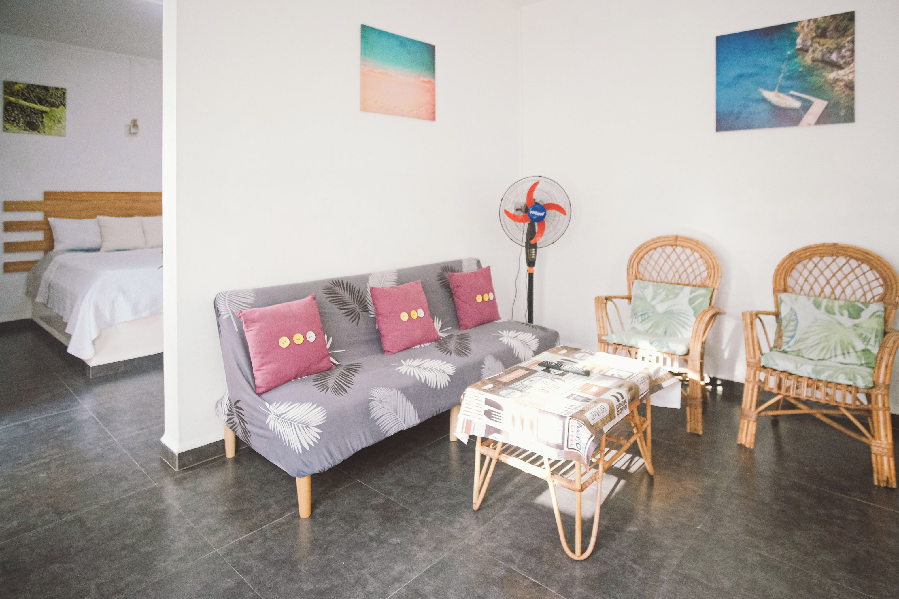 A modern room with a grey sofa, two wicker chairs, a small table, and a standing fan.