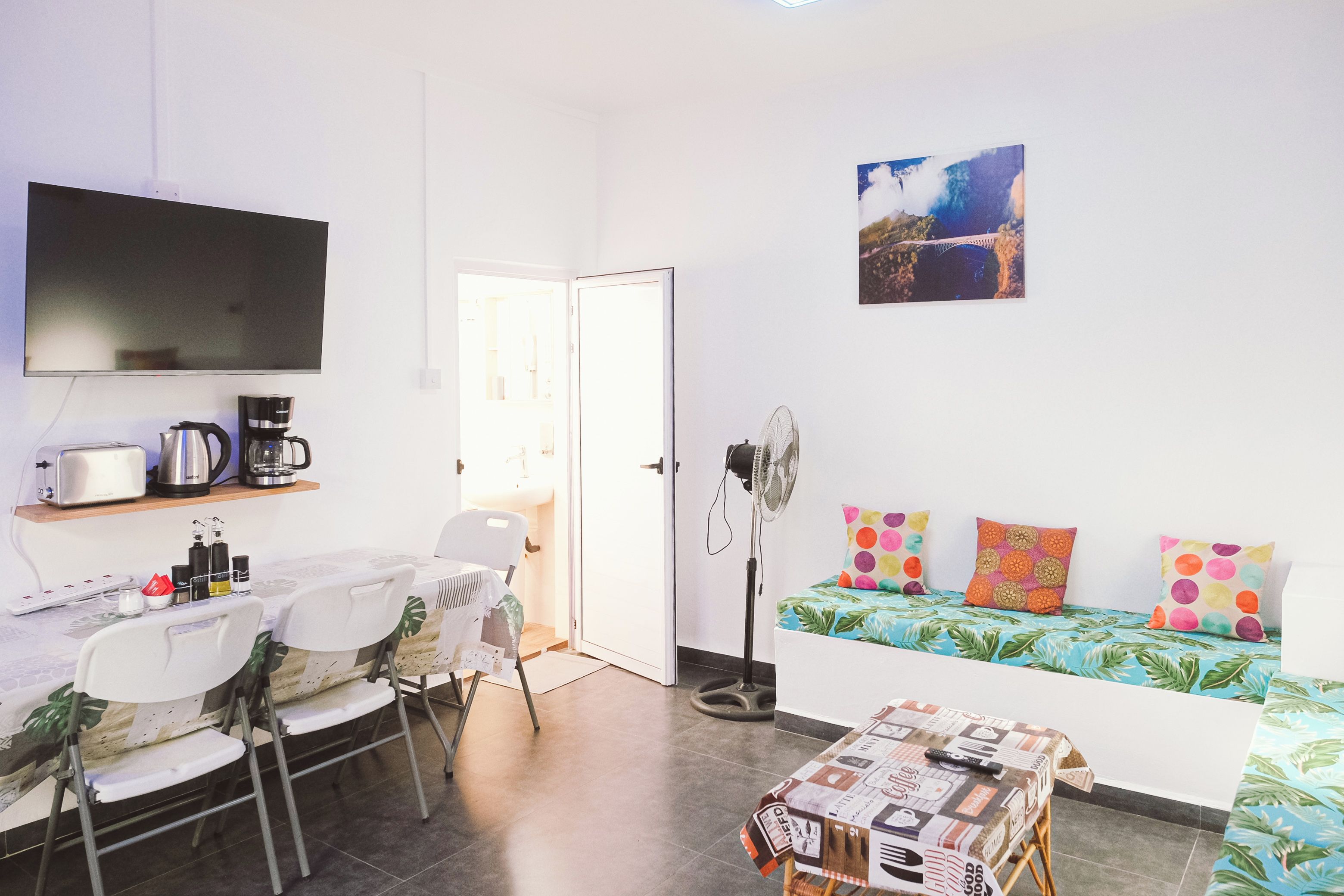 Een ruimtelijk ingerichte leefruimte met kitchenette, een eethoek, een bank met kleurrijke kussens en een aan de muur bevestigde televisie.