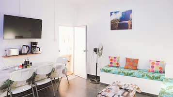 Een ruimtelijk ingerichte leefruimte met kitchenette, een eethoek, een bank met kleurrijke kussens en een aan de muur bevestigde televisie.