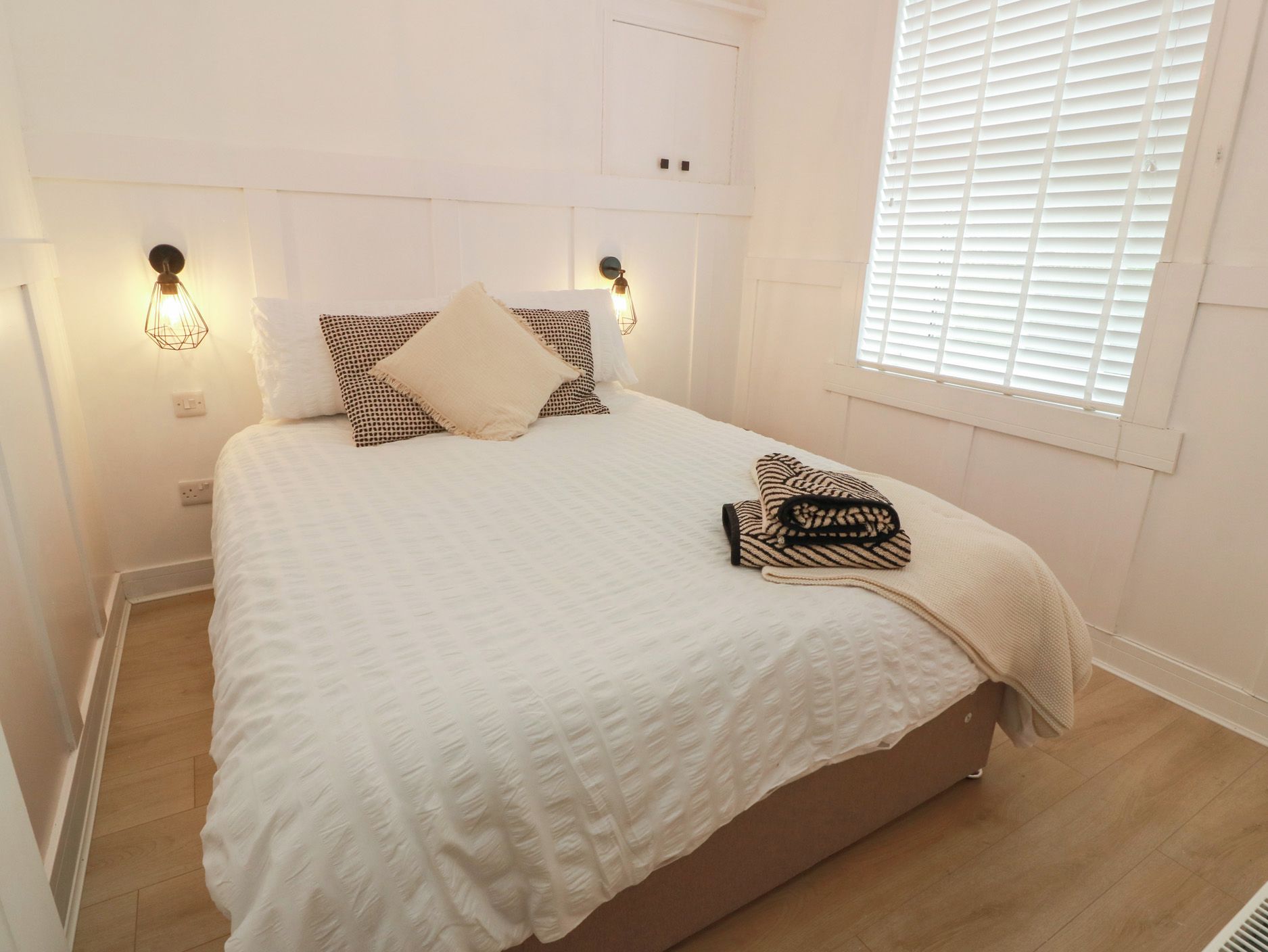 A bedroom with a large bed, two wall-mounted light fixtures, and a window with blinds.