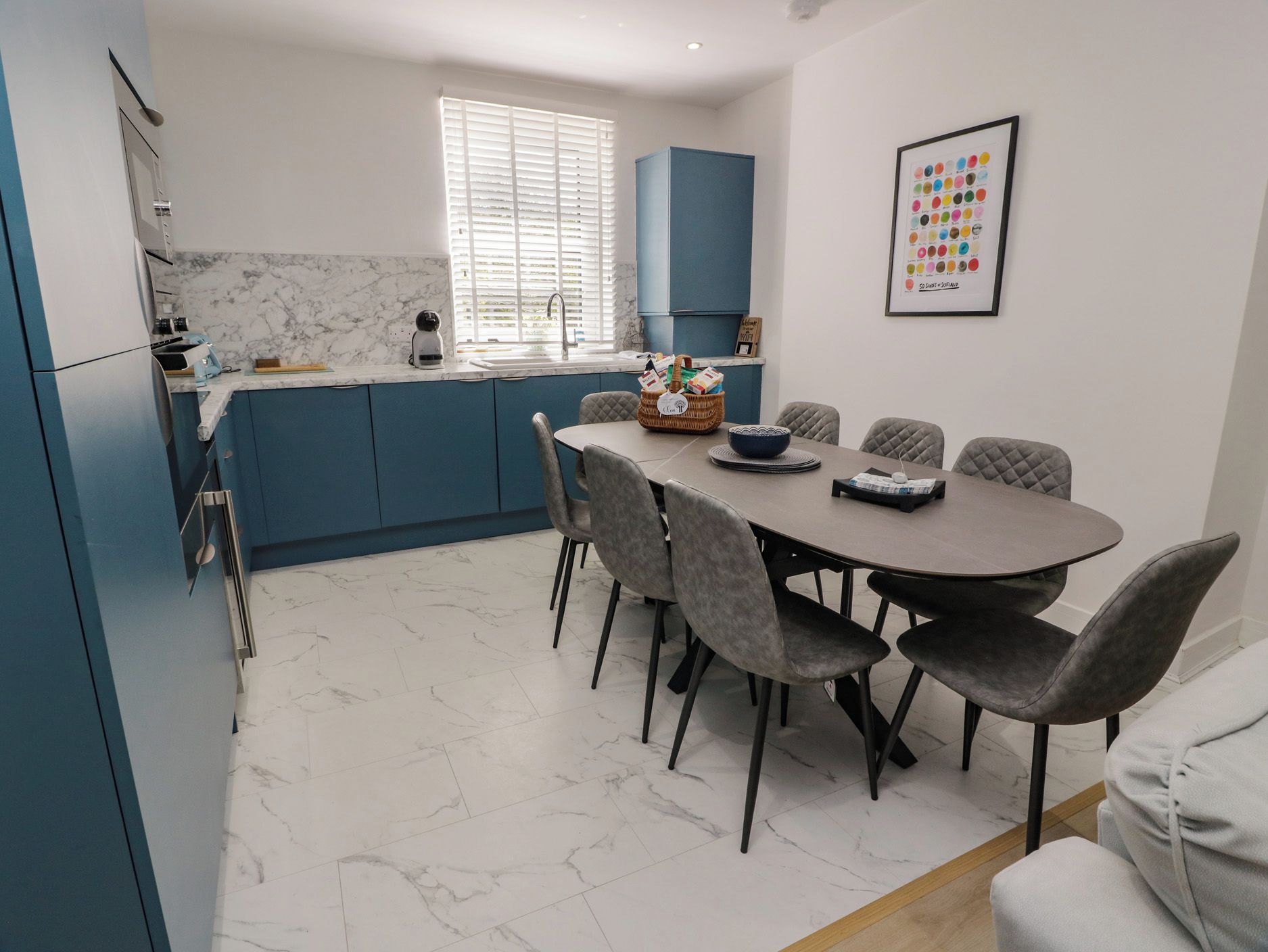 A modern kitchen with blue cabinets, a marble countertop, and a dining area with grey upholstered chairs.