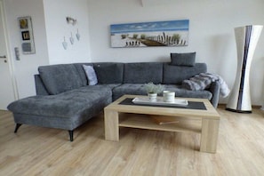 A modern living room with a grey sectional sofa, a wooden coffee table, and a painting of a beach scene. - Holiday apartment with 2 bedrooms (Horumersiel)