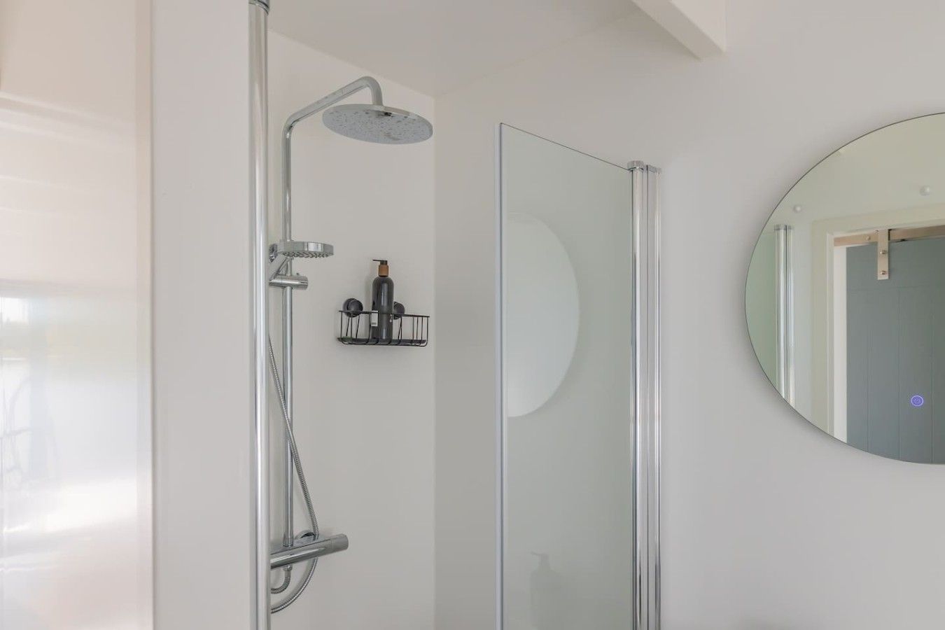Un baño moderno con cabina de ducha de vidrio, un espejo redondo y un pequeño estante con artículos de aseo.