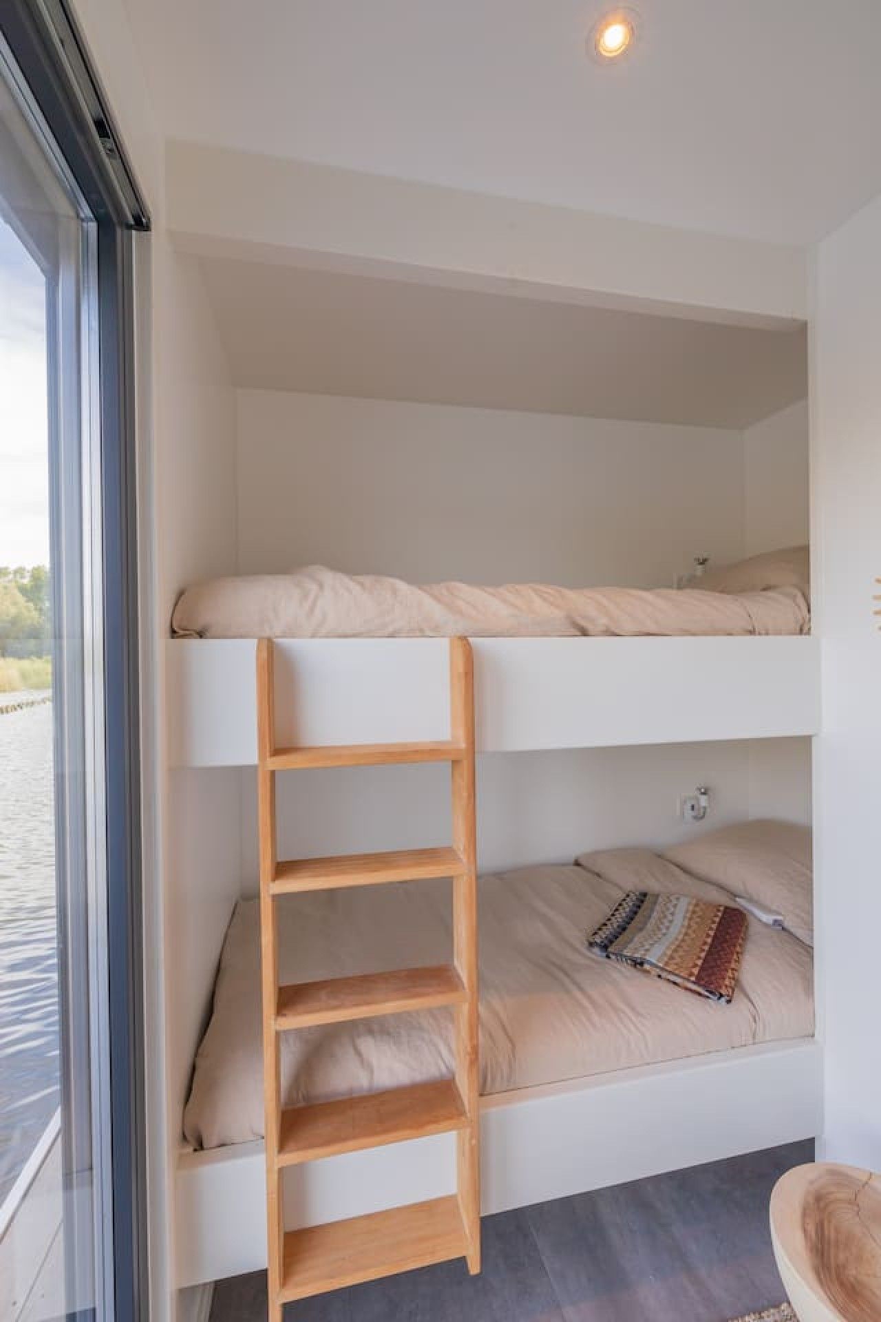 A bunk bed with a wooden ladder, a window with a view of a body of water, and a small shelf unit.
