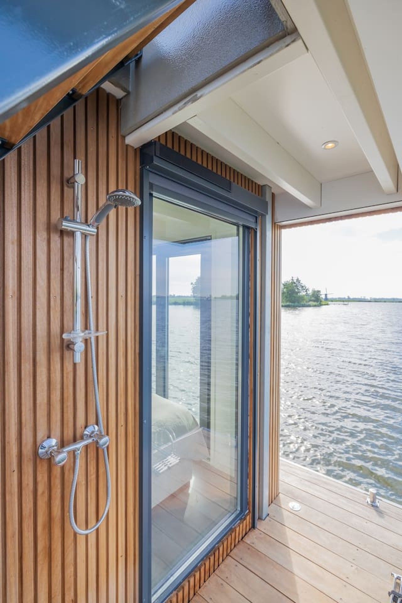 A modern bathroom with a shower, wooden paneling, and a glass door leading to a balcony with a view of a body of water.