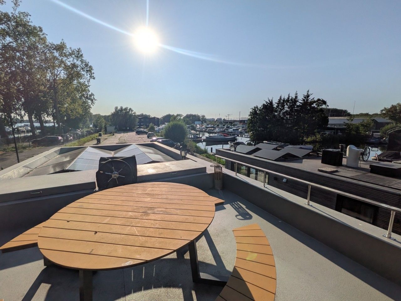 Una terraza en la azotea con una mesa redonda y bancos, con vistas a un aparcamiento y árboles.