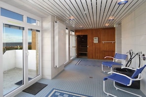 A corridor with a glass door leading to a balcony, a wooden wall, and a seating area with blue chairs and a black bathtub. - Luca Panorama Apt. 227, 1st floor (Cuxhaven)