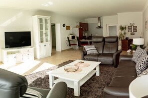 A living room with a television, a sofa, a coffee table, and a kitchen area in the background. - Luca Panorama Apt. 227, 1st floor (Cuxhaven)
