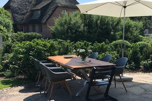 Ein Essbereich im Freien mit einem Holztisch und -stühlen, einem weißen Sonnenschirm und einem Haus mit einem Strohdach im Hintergrund.