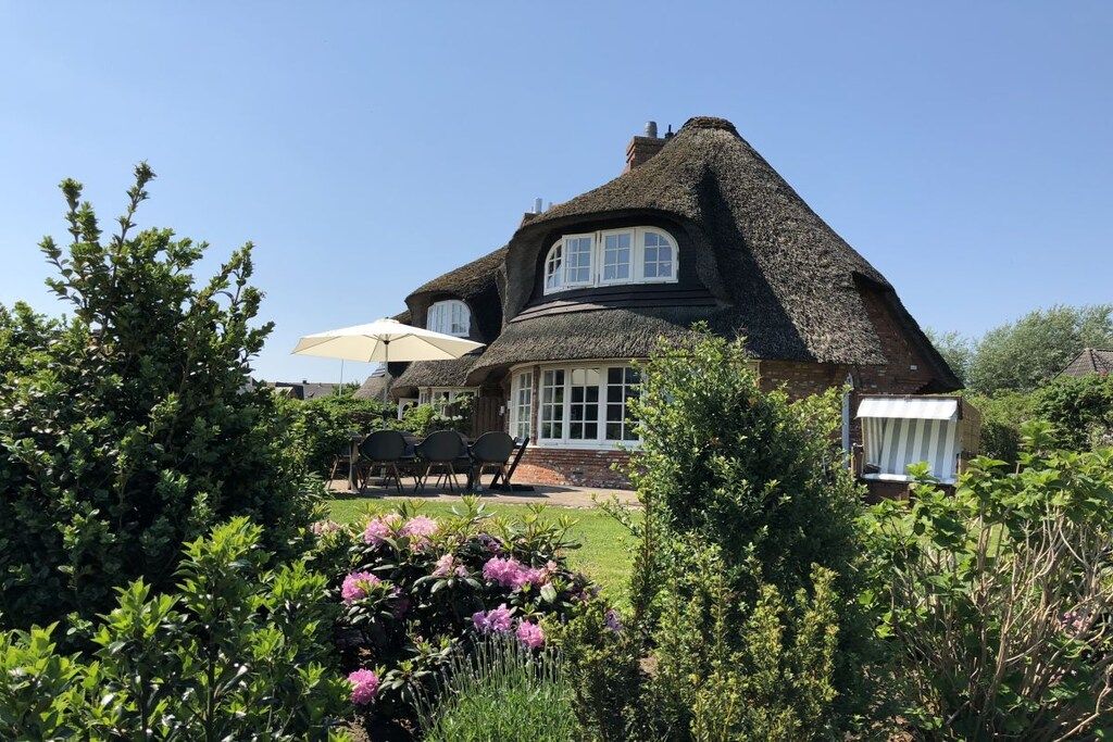 A house with a thatched roof, a garden with flowers, and a patio with chairs and an umbrella.