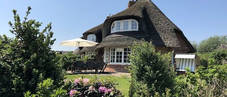 A house with a thatched roof, a garden with flowers, and a patio with chairs and an umbrella.