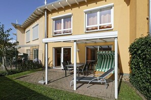 Ein zweistöckiges Haus mit einer überdachten Terrasse, Gartenmöbeln und einem grün gestreiften Sofa.