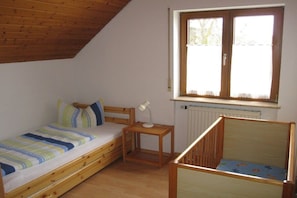 A small, single-room accommodation with a sloped ceiling, a bed with a striped bedspread, a small table with a lamp, a crib, and a window with a lace curtain. - Bernhard Winery Type B (Daisendorf)