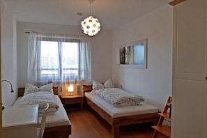 Una habitación con dos camas de madera, un escritorio, una silla, una ventana con cortinas, una lámpara colgante y un cuadro en la pared.