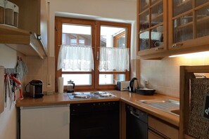 A kitchen with a stove, oven, and sink, wooden cabinets, and a window with lace curtains. - Holiday apartment with 3 bedrooms (Rettenberg)