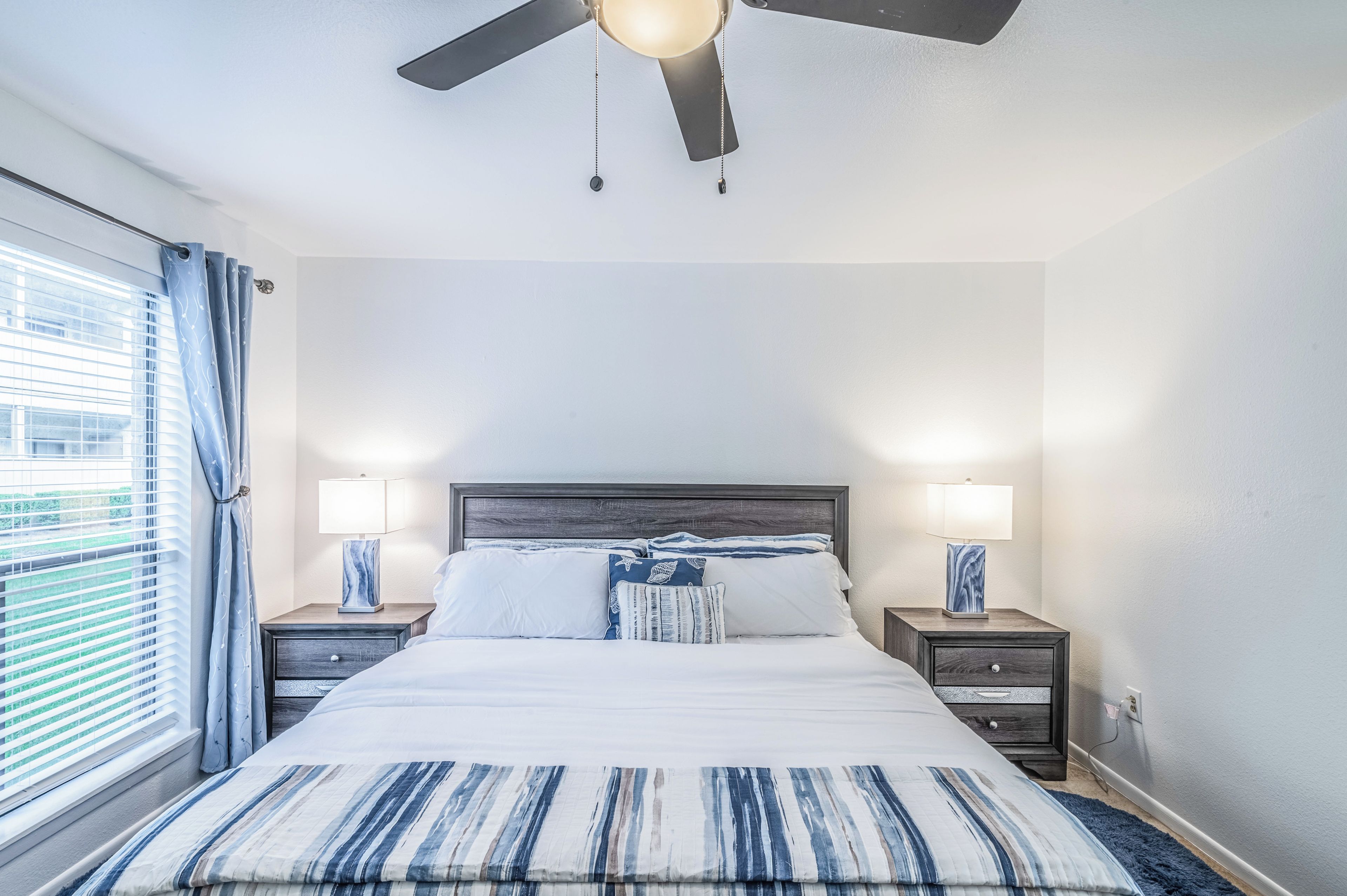 A bedroom with a bed, two bedside tables, a ceiling fan, and a window with curtains.
