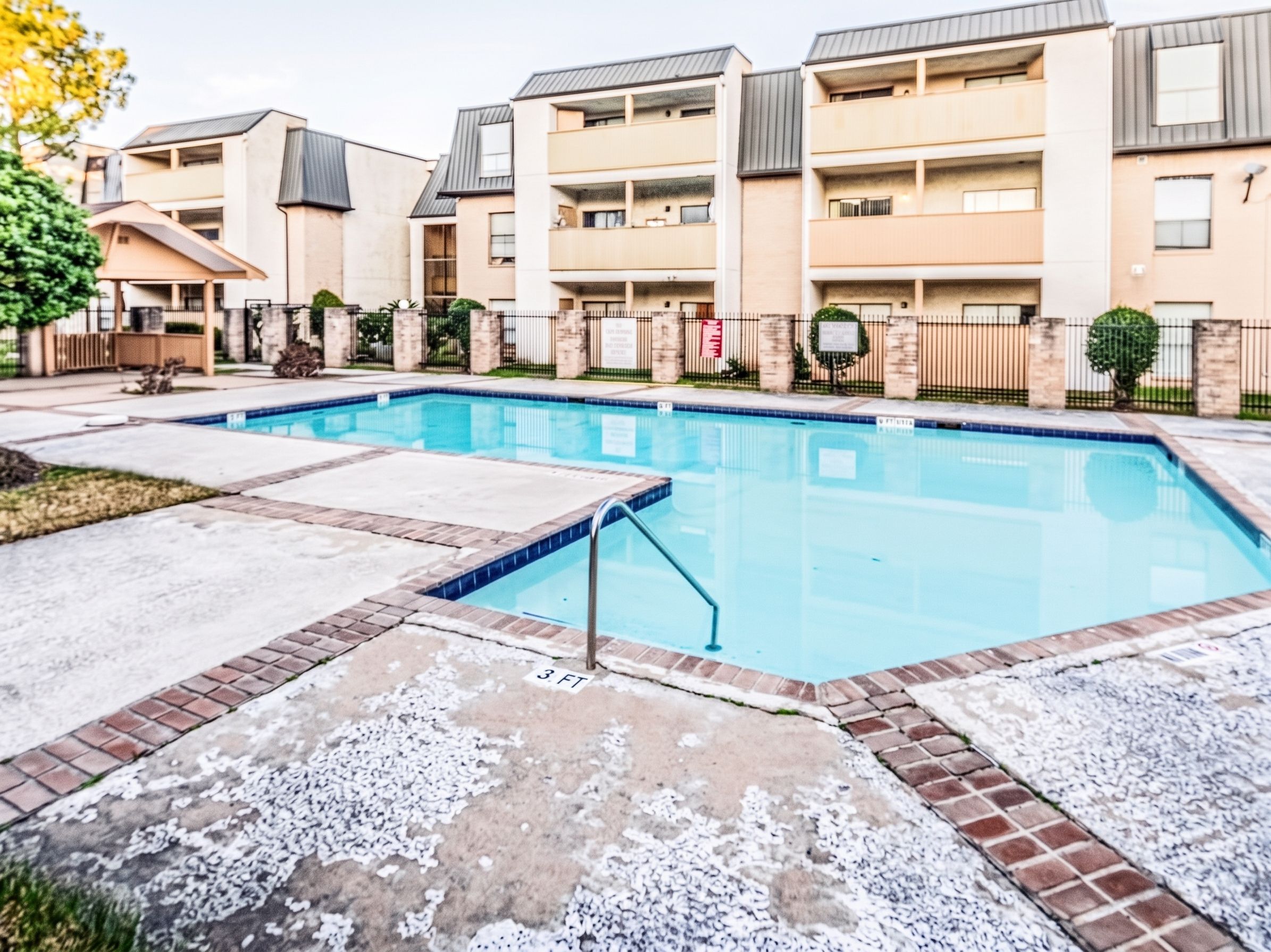 A swimming pool with a 3 ft depth marker, surrounded by a brick border and a metal fence.