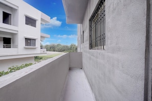 Uma varanda estreita com chão de azulejos brancos, limitada por um muro baixo e um edifício com janelas gradeadas.