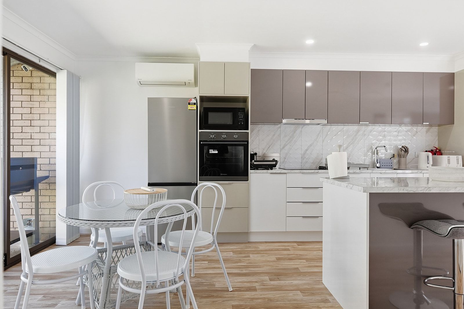 Une cuisine moderne équipée d’un four encastré, d’un micro-ondes et d’un réfrigérateur. On y trouve également un coin repas avec une table en verre et des chaises blanches.
