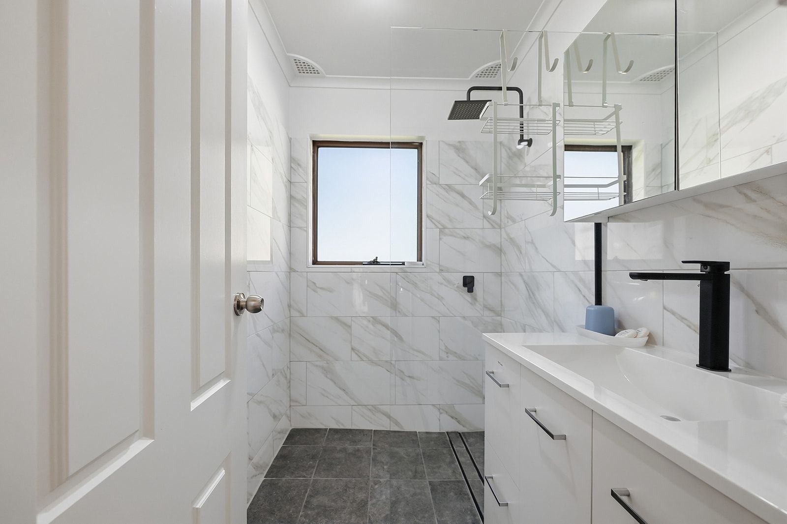 Un baño moderno con pared de mármol blanco, una ventana, una ducha y un lavabo con grifo negro.
