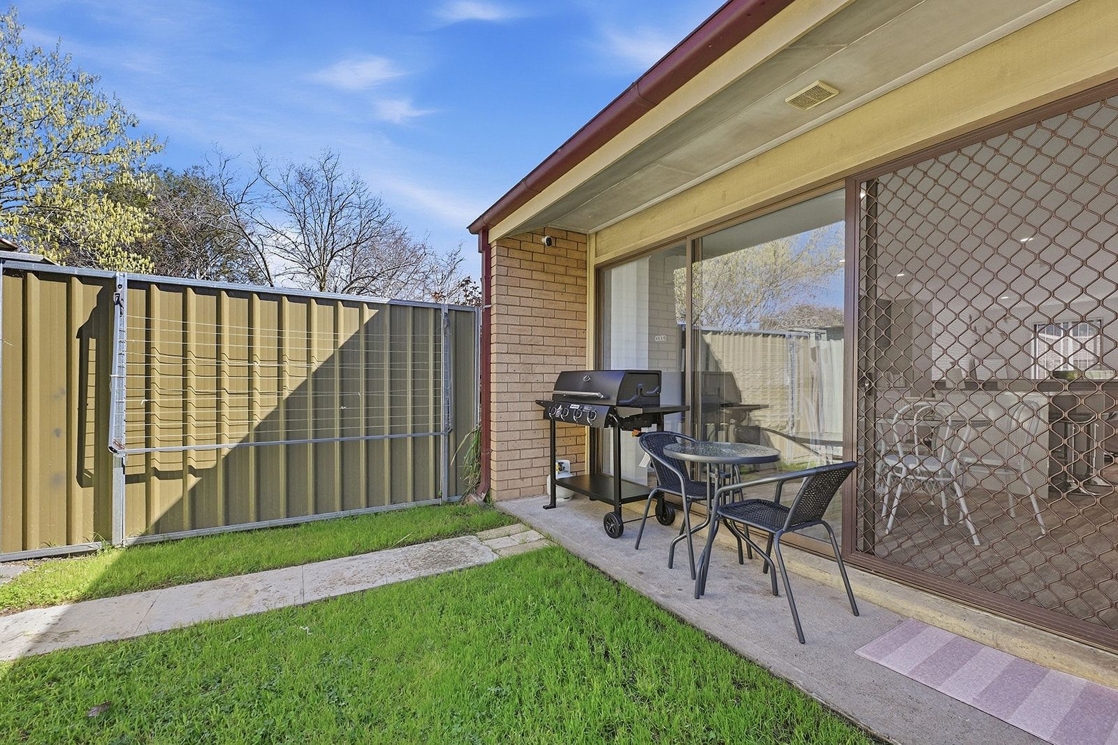 Un jardin avec un barbecue, une table et des chaises, le tout délimité par une clôture et accessible par une porte-fenêtre coulissante.