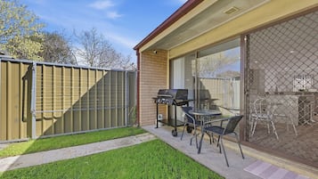 Un jardin avec un barbecue, une table et des chaises, le tout délimité par une clôture et accessible par une porte-fenêtre coulissante.