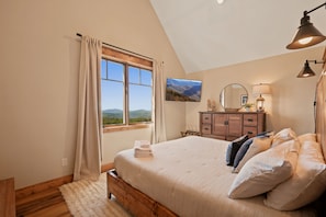 A bedroom with a large bed, a wooden dresser, a TV, and a view of mountains through the window. - Evenlight Retreat: Sunset Views, Hot Tub, Game Room, A/C | Eagle's Nest (Elk Park)