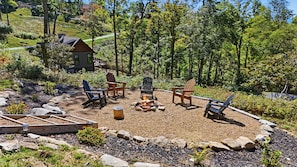 A circular outdoor seating area with Adirondack chairs and a fire pit, surrounded by a gravel and rock border, with a view of a house and forest in the background. - Evenlight Retreat: Sunset Views, Hot Tub, Game Room, A/C | Eagle's Nest (Elk Park)
