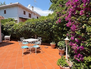 Eine Terrasse mit Tisch und Stühlen, umgeben von Blumentöpfen und einem Hecken.