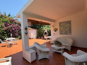 Un patio con mobiliario de mimbre, zona de barbacoa y jardín con flores púrpuras.