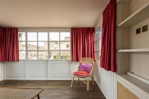 A room with a large window, a wicker chair with a cushion, and a white wall with a shelf. - Trastevere Luxury Panoramic Terrace (Roma)