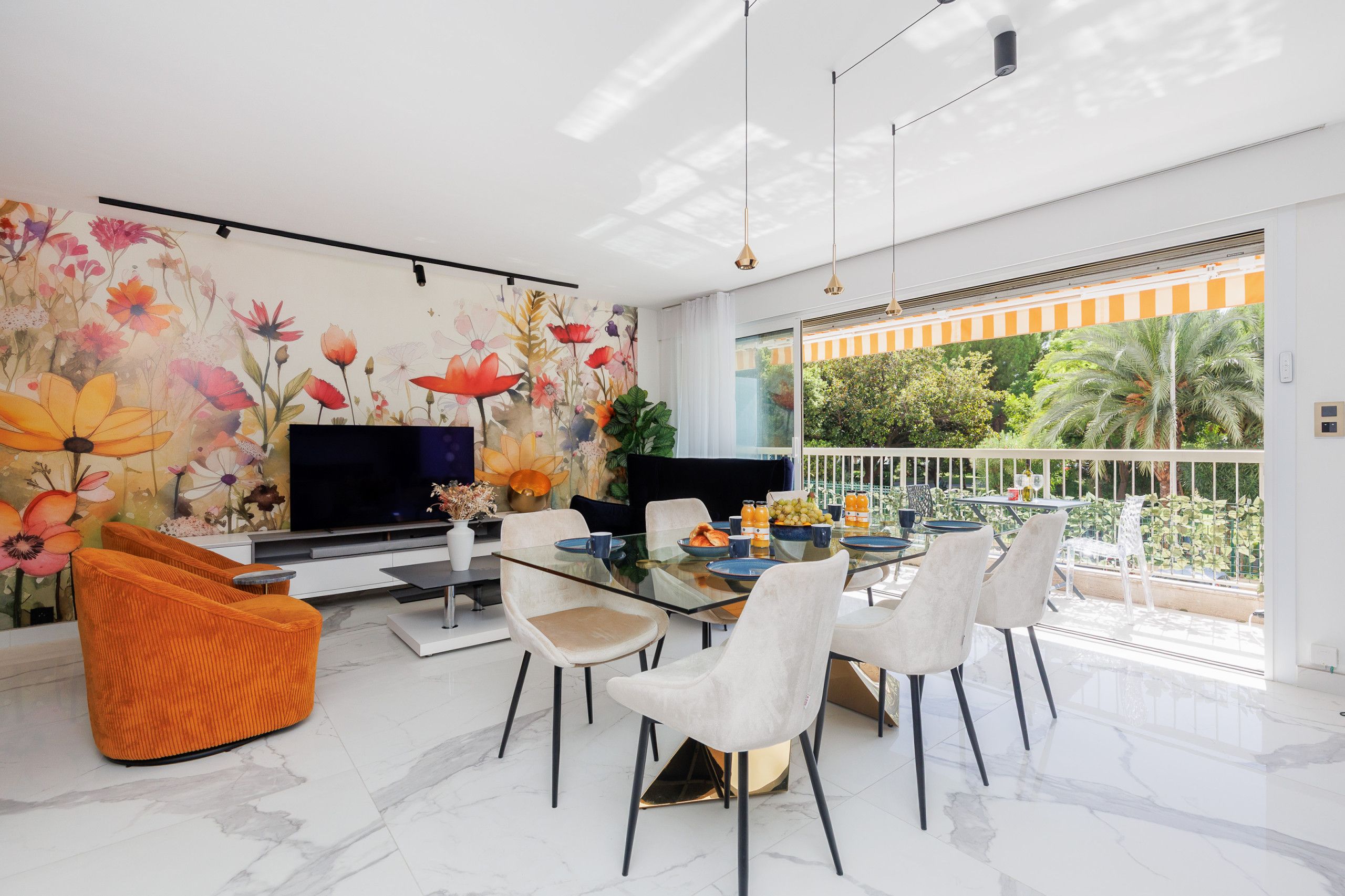 A modern living room with a flat-screen TV, a dining area with a glass table, and a balcony with a view of greenery.