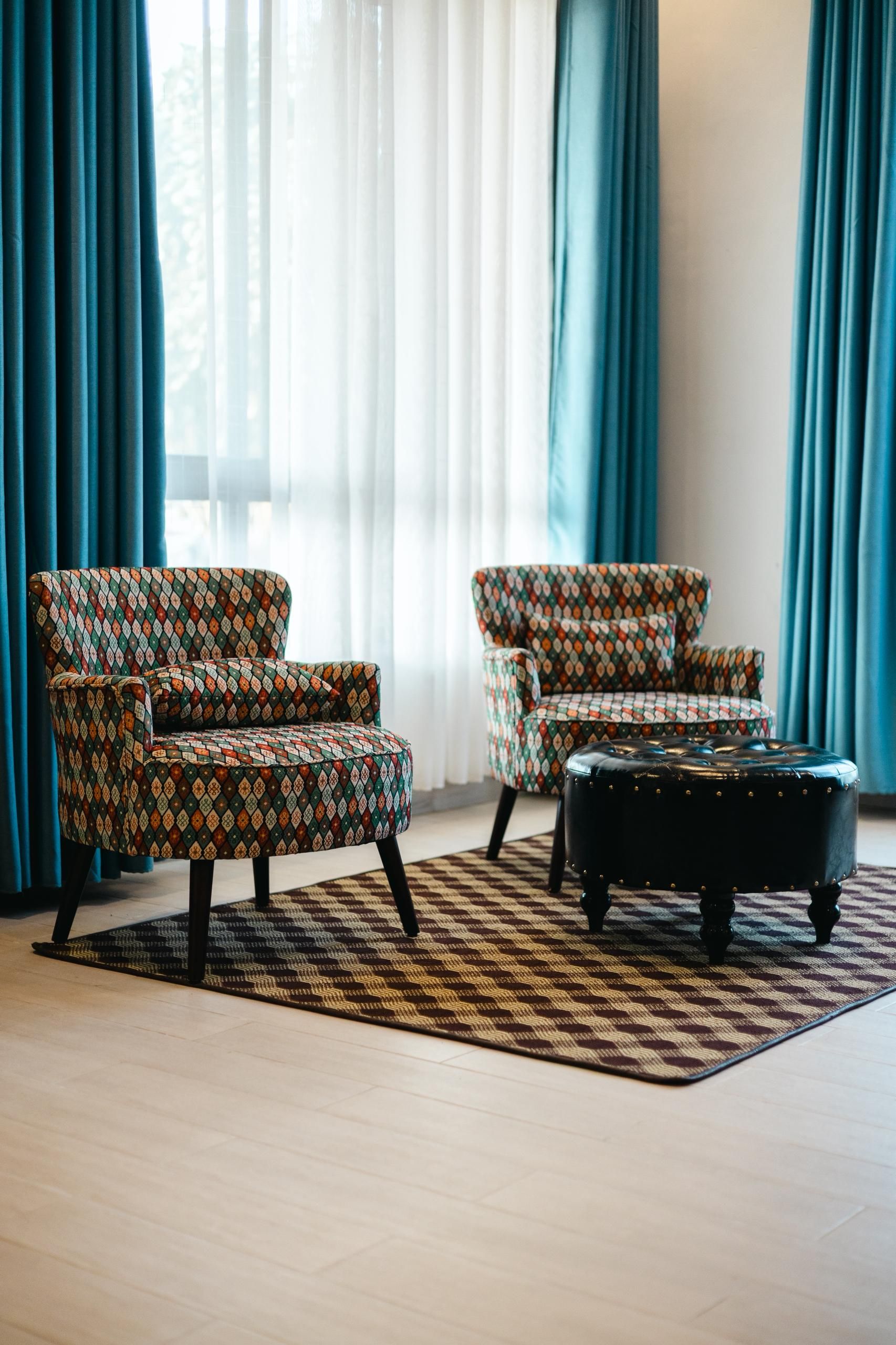 A living room with two patterned armchairs and a black ottoman on a patterned rug.
