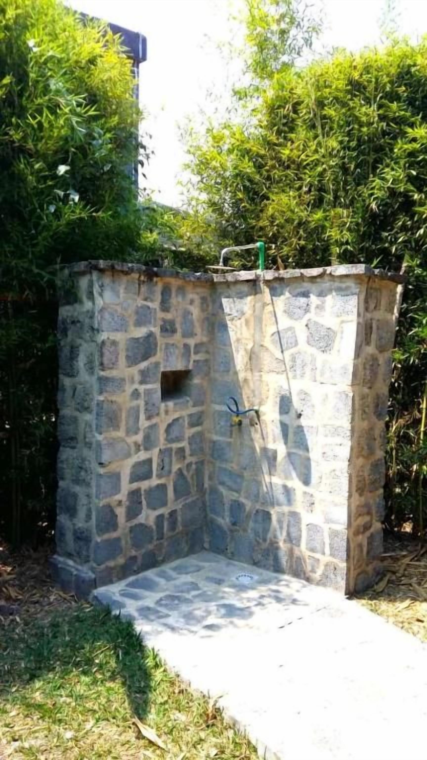 A stone-built outdoor shower area with a concrete floor, surrounded by greenery.