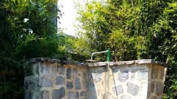 A stone-built outdoor shower area with a concrete floor, surrounded by greenery.
