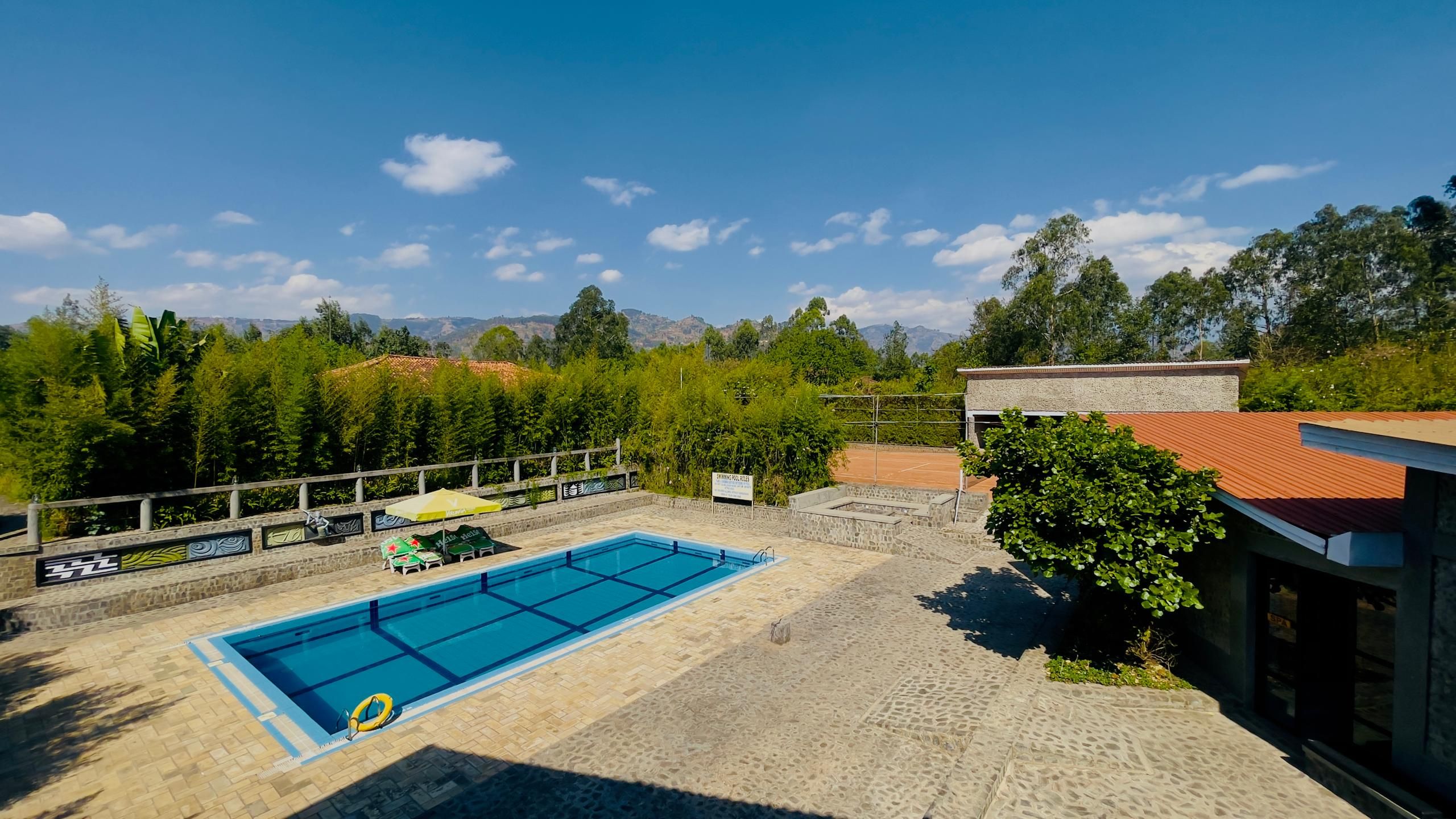 A swimming pool with a tiled deck, surrounded by a fence and trees, with mountains in the background.
