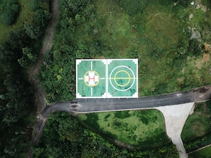 An aerial view of a basketball court with a hospital symbol, surrounded by dense greenery and a bridge.